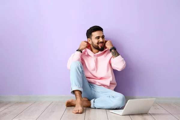 Handsome barefoot man with laptop sitting near lilac wall