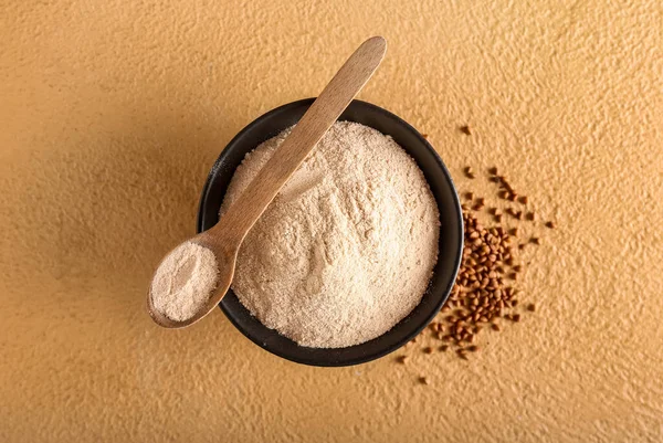 Bej arka planda bir kase un, kaşık ve karabuğday taneleri.
