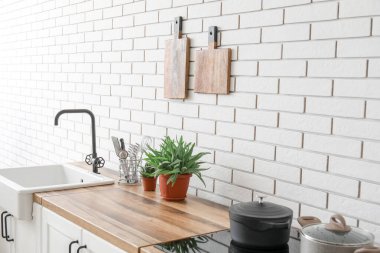 Mutfak tezgahları beyaz tuğla duvarın yanında lavabo, ev bitkileri ve çatal bıçaklarla dolu.