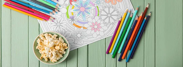 Composition with coloring picture, pencils and popcorn on color wooden table, top view