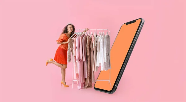 Stylish young woman with stylish clothes on hanger and modern mobile phone on pink background