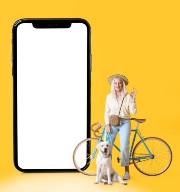 Sarı arka planda köpeği, bisikleti ve modern cep telefonu olan şık, olgun bir kadın.