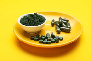 Bir kase spirulina tozu ve sarı arka planda haplar.