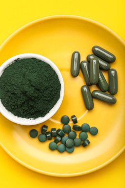 Bir kase spirulina tozu ve sarı arka planda haplar.