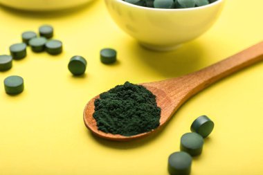 Sarı arka planda bir kaşık spirulina tozu ve haplar, yakın plan.