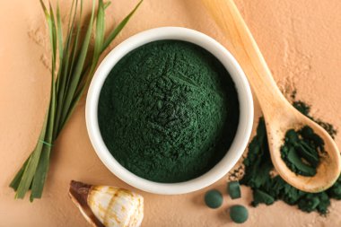 Bir kase spirulina tozu, kaşık, hap, deniz kabuğu ve bej arka planda yapraklar.