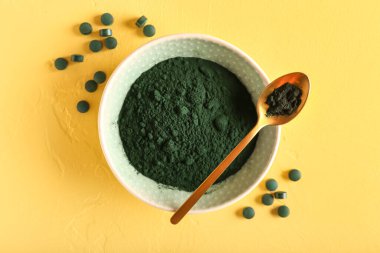Sarı arka planda bir kase spirulina tozu, kaşık ve haplar.