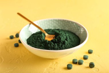 Sarı arka planda bir kase spirulina tozu, kaşık ve haplar.
