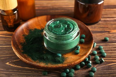 Tahta arka planda bir kavanoz spirulina yüz maskesi, toz ve hapla kaplanmış.