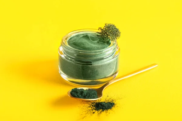 Jar of spirulina facial mask and spoon with powder on yellow background