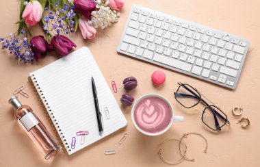 Bir fincan pembe kibrit çay, bilgisayar klavyesi, defter, çiçekler ve arka planda kadın aksesuarları.