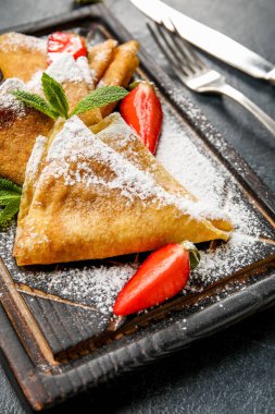 Koyu arkaplanda ince krep, çilek ve şeker tozu olan tahta, yakın plan.