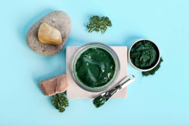 Mavi arka planda spirulina yüz maskesi, taşlar ve barut kavanozu olan kompozisyon