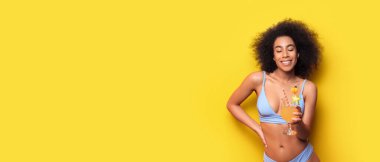 Bikinili güzel Afro-Amerikalı kadın ve sarı arkaplanlı kokteyl. Mesaj için yeri var.