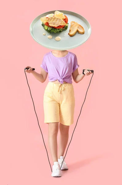 Sporty young woman with skipping rope and plate of tasty Caesar salad instead of her head on pink background