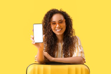 Sarı arka planda cep telefonu ve bavulu olan Afro-Amerikalı genç bir kadın.