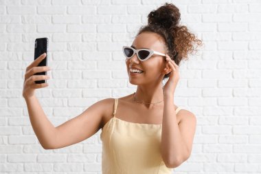 Beyaz tuğlalı arka planda cep telefonuyla selfie çeken Afro-Amerikalı kadın turist.