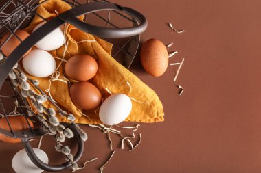 Overturned basket with different chicken eggs and pussy willow branches on color background