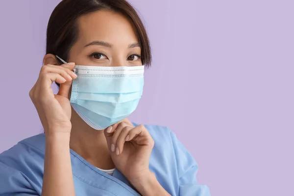 Female Asian nurse putting on medical mask against color background