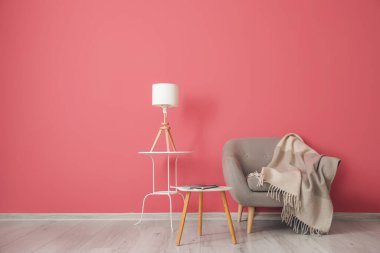 Comfortable armchair with plaid, tables and lamp near pink wall in room