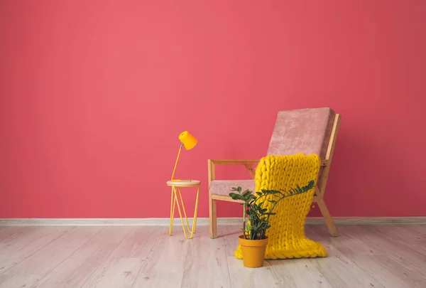 Comfortable armchair with plaid, houseplant, table and lamp near pink wall in room