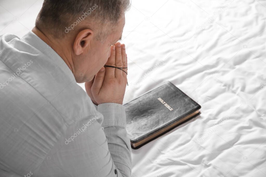 Praying mature man with Holy Bible and cross on bed