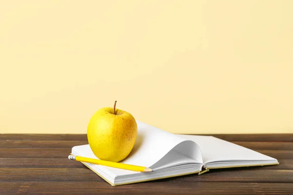 School book with apple and pencil on table against color background
