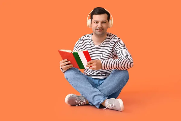 Young man with headphones and book on orange background. Concept of studying Italian language