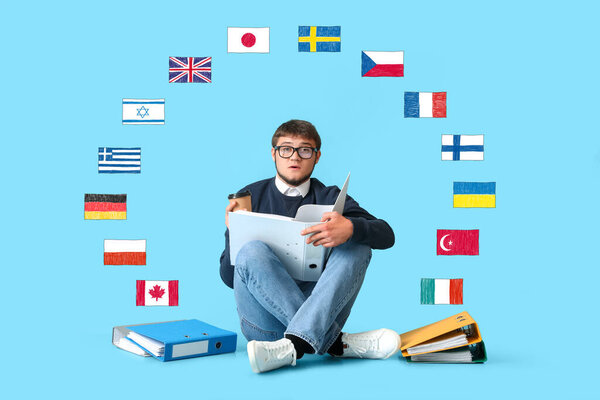 Young male student with many folders and different flags on light blue background. Studying of foreign languages