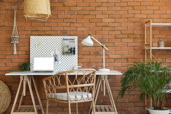 Modern workplace with peg board, laptop and lamp near brick wall