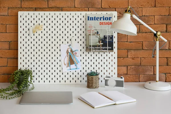 Modern workplace with peg board, laptop and lamp near brick wall