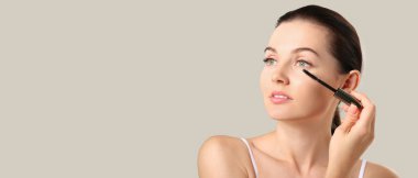 Gri arkaplan üzerine rimel süren genç ve güzel bir kadın.