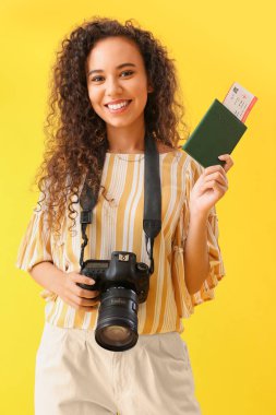 Sarı arka planda pasaport ve fotoğraf makinesi olan genç bir Afro-Amerikalı kadın.