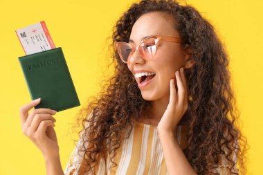 Sarı arka planda pasaportu ve bileti olan mutlu bir Afro-Amerikalı kadın.