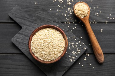 Koyu ahşap zemin üzerinde kase ve kaşık dolusu susam tohumu olan tahta.