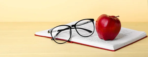 School book with apple and eyeglasses on table against color background