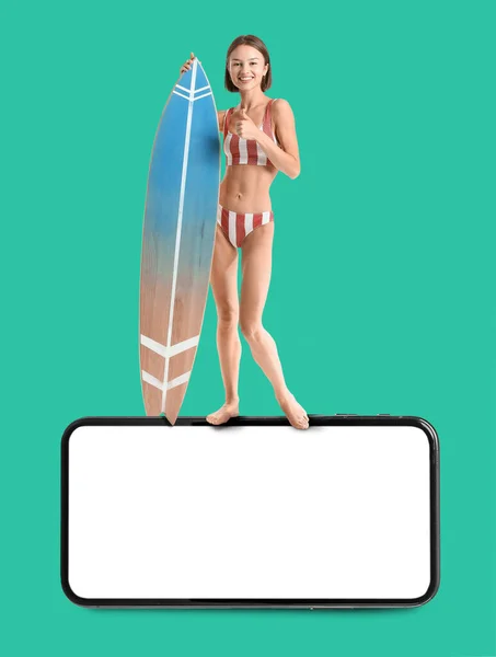 Young woman with surfboard and big mobile phone with blank screen on color background