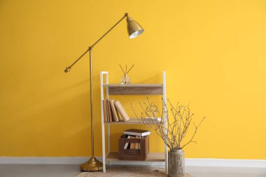 Wooden bookcase, vase with tree branches and lamp near yellow wall