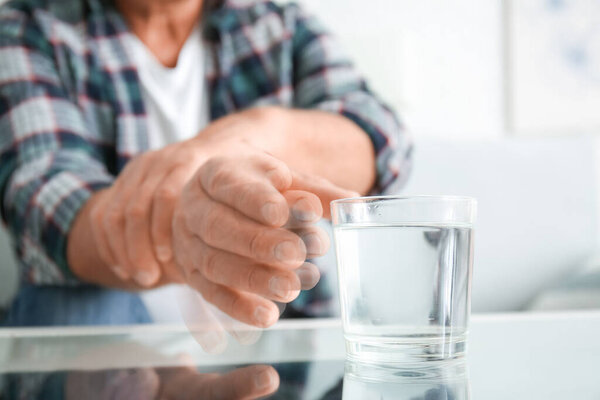 Senior man with Parkinson syndrome taking glass of water from table, closeup