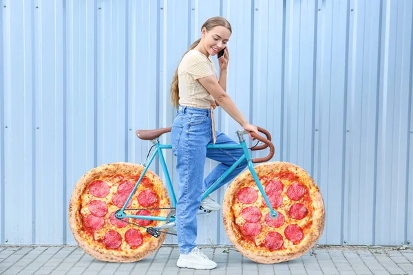 Beautiful teenage girl with mobile phone and bicycle with pizza pepperoni instead of wheels outdoors