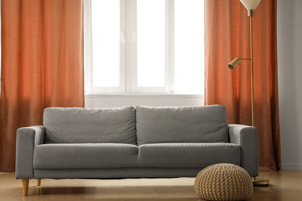 Серый диван, пуф и стандартная лампа возле красных занавесок в номере