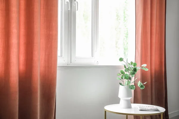 Vase with eucalyptus branches on table near red curtains in room