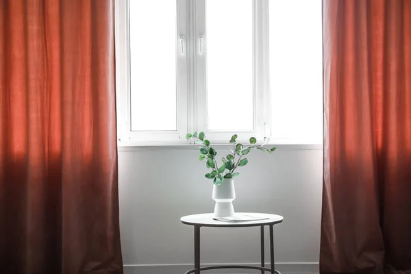 Vase with eucalyptus branches on table near red curtains in room