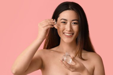 Young Asian woman with cosmetic serum on pink background