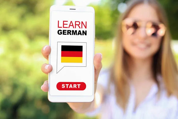Woman holding mobile phone with text LEARN GERMAN on screen outdoors, closeup
