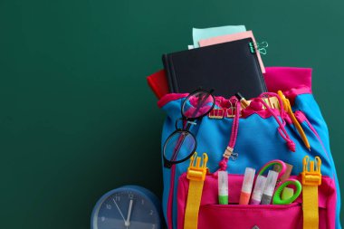 Yeşil karatahtanın yanında kırtasiye malzemesi ve alarm saati olan okul çantası