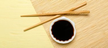 Bir kase soya sosu, yemek çubukları ve sarı ahşap arka planda bambu hasırı.