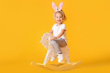 Cute little girl in bunny ears with rocking horse on yellow background