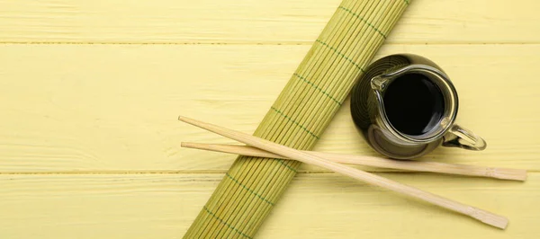 Jug of soy sauce, chopsticks and bamboo mat on yellow wooden background with space for text, top view