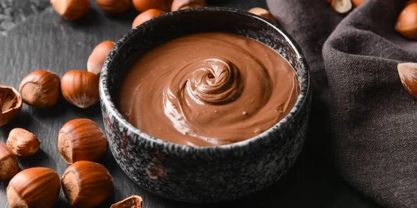 Bowl with tasty chocolate paste and hazelnuts on dark background, closeup
